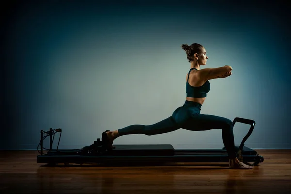 Chica joven haciendo ejercicios de pilates con una cama reformadora. Hermoso entrenador de fitness delgado sobre fondo gris reformador, bajo llave, luz de arte. Concepto Fitness — Foto de Stock