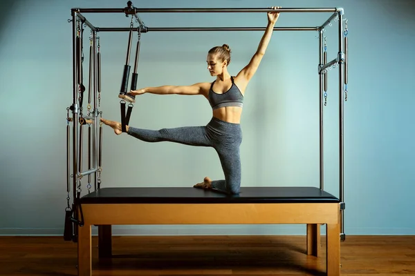 Giovane ragazza che fa esercizi di pilates con un letto riformatore. Bellissimo allenatore di fitness sottile su sfondo grigio riformatore, basso profilo, luce d'arte. Concetto fitness — Foto Stock