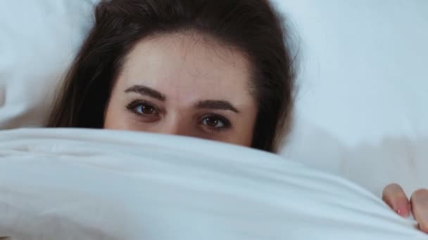 Vista superior de una joven alegre escondida bajo una suave manta blanca, modelo más tamaño divirtiéndose en la cama, mirando a la cámara, disfrutando de buenos días, descansando en la cama. Vista desde arriba. Copiar espacio — Vídeo de stock