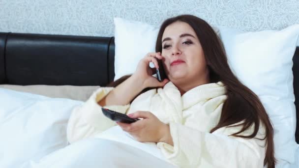 Девушка разговаривает по телефону, когда лежит в кроватке. Поделитесь новостями во время карантина, оставайтесь дома. Модель плюс размер. Портрет крупным планом домохозяйки — стоковое видео