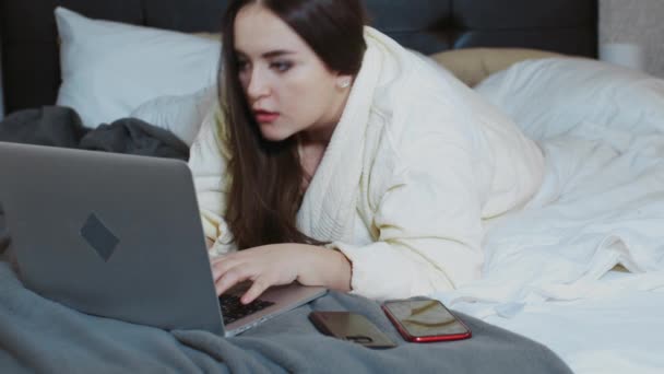 Vrouw in badjas liggend op het bed met een laptop en smartphones voor het werk. Freelance, thuis werken, buiten het kantoor werken. Moderne technologieën en communicatiemiddelen — Stockvideo