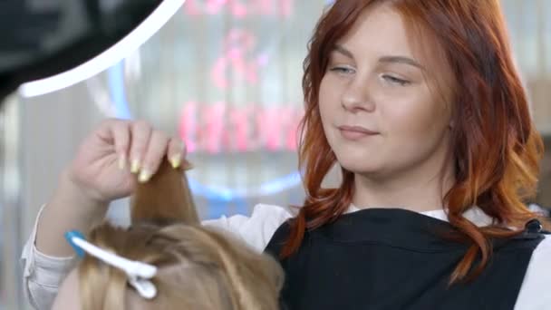 クライアントの髪で働く美しい赤い髪のマスターは、カーリングアイロンを使用して巻き毛をしています。美容室で長いブロンドの髪のためのカールヘアを作成します。. — ストック動画
