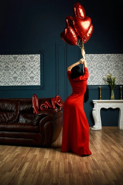 Sexy woman wearing in red dress standing near sofa and holding heart-shaped balloons and a love sign and looks very sensually — 스톡 사진