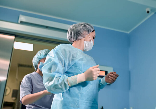 Команда хирургов готовится к операции. Хирурги носят стерильную одежду перед операцией с помощью медсестер, стерильных халатов, перчаток, масок, в отделении интенсивной терапии.