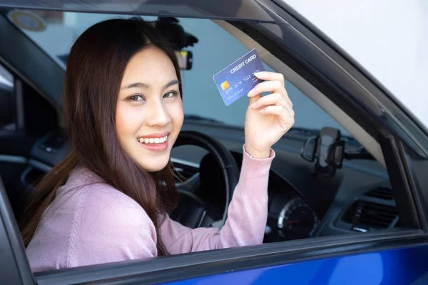 Mulher asiática bonita feliz sentada dentro do carro novo azul e mostrando o pagamento do cartão de crédito para o petróleo, pagar um pneu, manutenção na garagem, fazer o pagamento para reabastecer o carro no posto de gasolina, financiamento automotivo — Fotografia de Stock