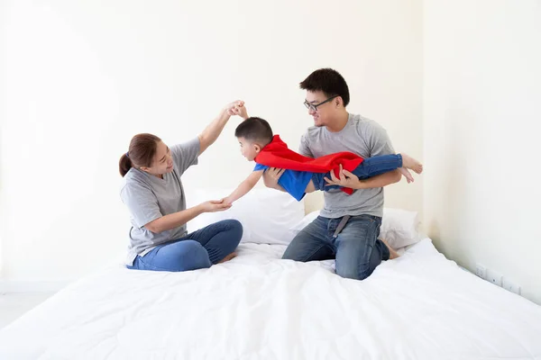 Pai asiático, mãe e filho estão jogando super-herói na cama no quarto. Família amigável se divertindo — Fotografia de Stock