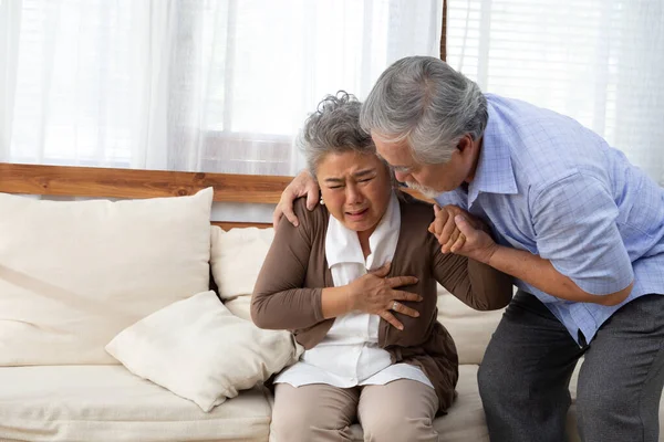 Asian senior woman with pain on heart on sofa in living room and take care by her husband, Health problem and feeling sick