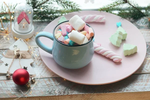 A cup with a drink and marshmallows in hands in gloves on the background of Christmas decor, illumination on a wooden windowsill, the concept of home comfort, winter holidays