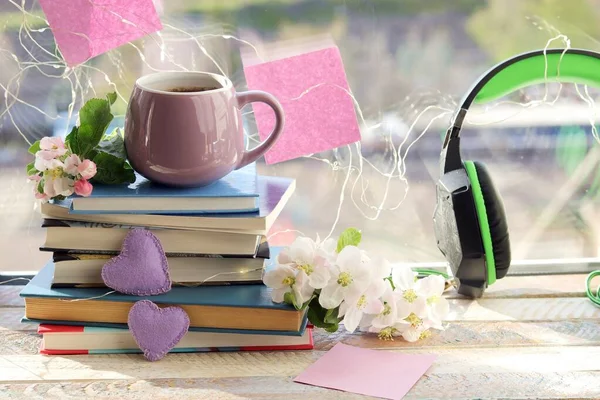 Bir fincan kahve, kalplerle süslenmiş bir yığın kitap, ahşap pencere eşiğinde elma çiçekleri, ev konforu.