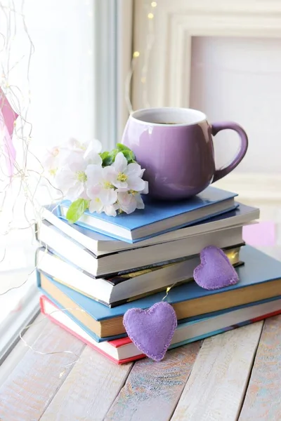 Cup of coffee, a stack of books decorated with hearts, apple flowers on a wooden windowsill, the concept of home comfort