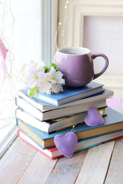 Bir fincan kahve, kalplerle süslenmiş bir yığın kitap, ahşap pencere eşiğinde elma çiçekleri, ev konforu.