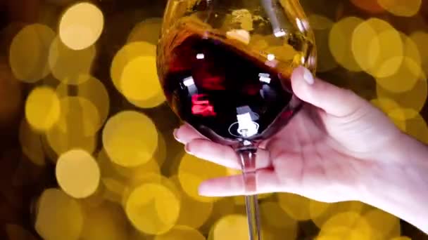 Female hand holds glass of red wine and moves on background of golden highlights — Stok video