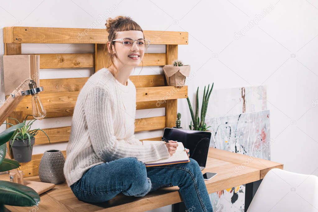 smiling girl sitting on table in office and writing something to notebook