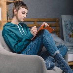 Girl sitting on armchair in bedroom and writing something to notebook