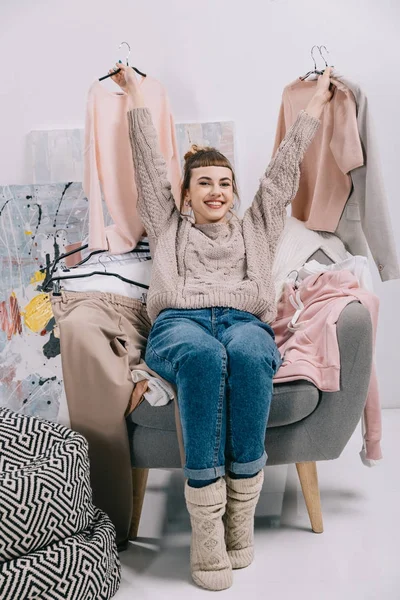 Chica sonriente sentada en un sillón y sosteniendo perchas con ropa en las manos - foto de stock