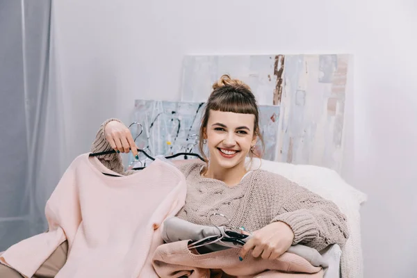 Chica sonriente sentada en sillón con ropa en perchas y mirando a la cámara - foto de stock
