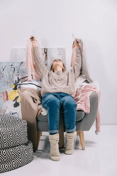 Girl lying on armchair and holding hangers with clothes in hands — Stock Photo