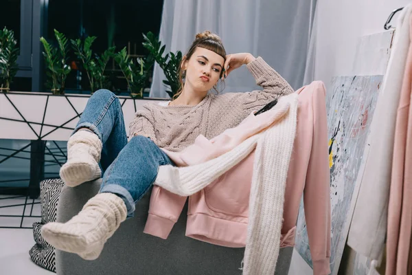 Девушка сидит на кресле и скептически смотрит на свитера на вешалках — стоковое фото