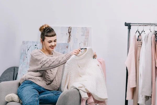 Chica sonriente sentada en el sillón y mirando suéter en las manos - foto de stock