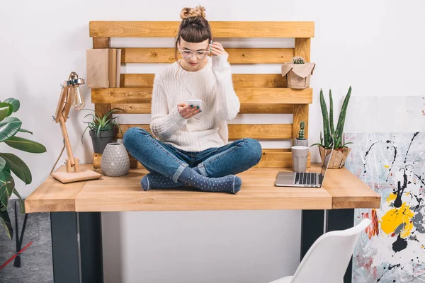 Girl sitting on table in office and looking at smartphone — Stock Photo