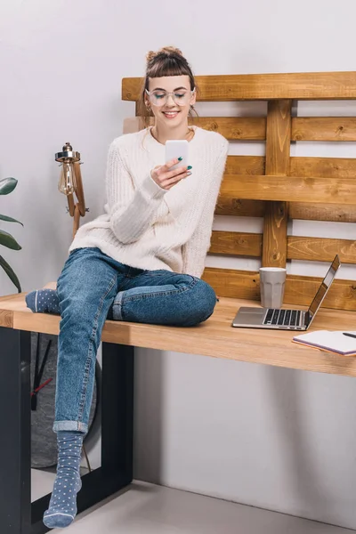 Lächelndes Mädchen, das im Büro auf dem Tisch sitzt und aufs Smartphone schaut — Stockfoto