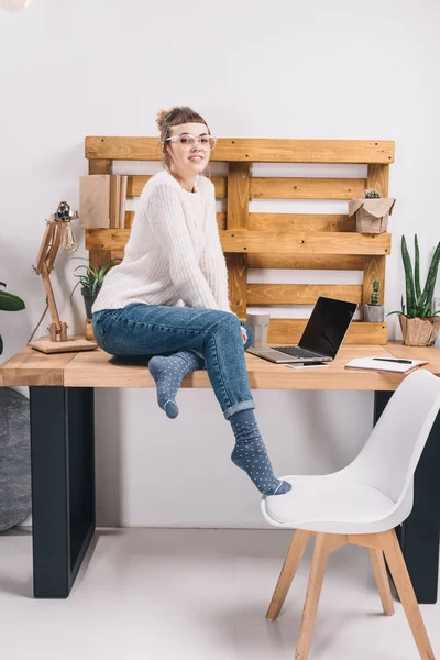 Chica sonriente sentada en la mesa en la oficina y mirando a la cámara - foto de stock