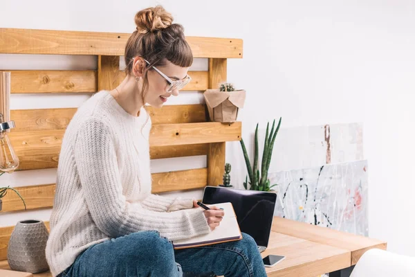 Chica sonriente sentada en la mesa en la oficina y escribiendo algo en el cuaderno - foto de stock