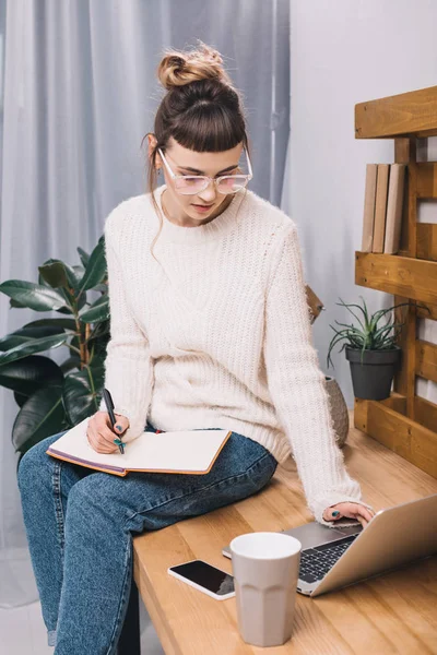 Mädchen sitzt auf Tisch im Büro, schreibt etwas in Notizbuch und benutzt Laptop — Stockfoto