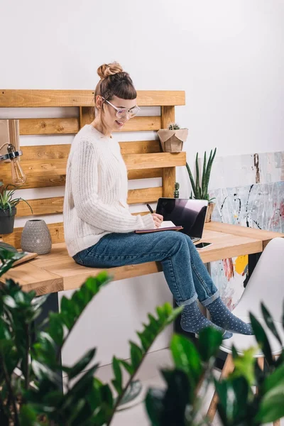 Smiling girl sitting on table in office and writing something to notebook — Stock Photo