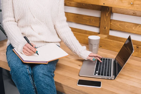 Cropped image of girl sitting on table in office, writing something to notebook and using laptop — Stock Photo