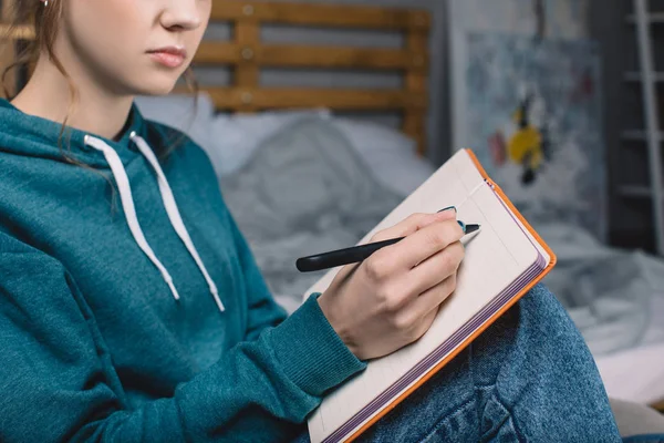 Abgeschnittenes Bild eines Mädchens, das auf einem Sessel im Schlafzimmer sitzt und etwas ins Notizbuch schreibt — Stockfoto