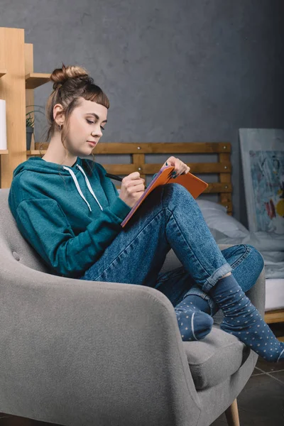 Girl sitting on armchair in bedroom and writing something to notebook — Stock Photo