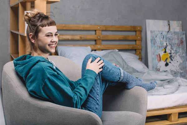 Chica sonriente sentada en el sillón en el dormitorio y mirando hacia otro lado - foto de stock