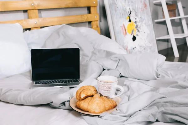 Cup of coffee and croissants on plate and open laptop on bed — Stock Photo