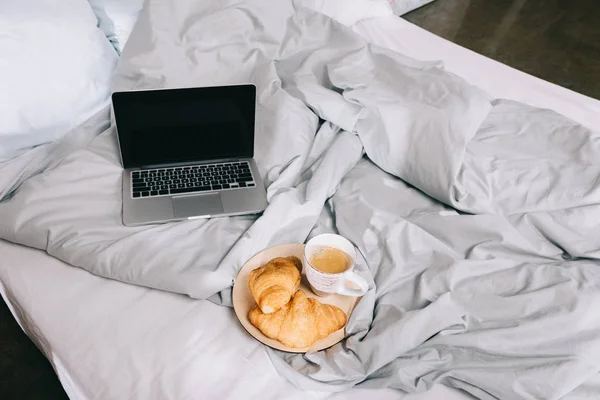 Tasse de café et croissants sur assiette et ordinateur portable ouvert sur lit — Photo de stock
