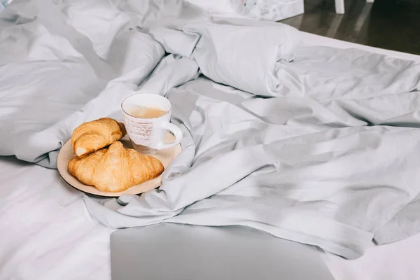 Tasse Kaffee und Croissants auf Teller und Laptop auf Bett — Stockfoto