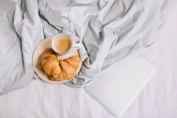 Vue du dessus de tasse de café et croissants sur assiette et ordinateur portable sur lit — Photo de stock