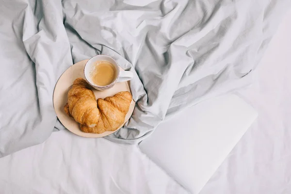 Vista superior de la taza de café y cruasanes en el plato y el ordenador portátil en la cama - foto de stock