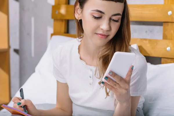 Chica feliz sentado en la cama por la mañana y mirando el teléfono inteligente - foto de stock