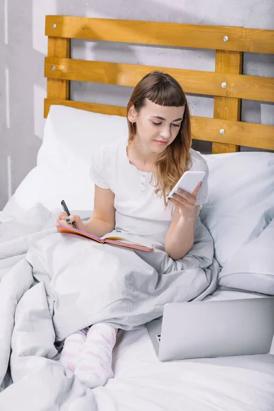 Happy girl sitting in bed in morning and looking at smartphone — Stock Photo