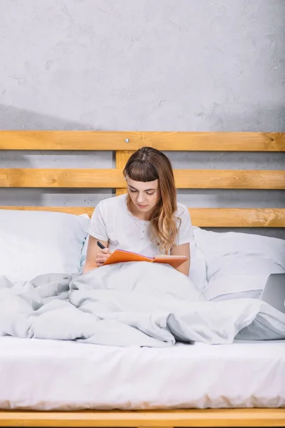 Menina sentada na cama e escrevendo algo para notebook — Fotografia de Stock
