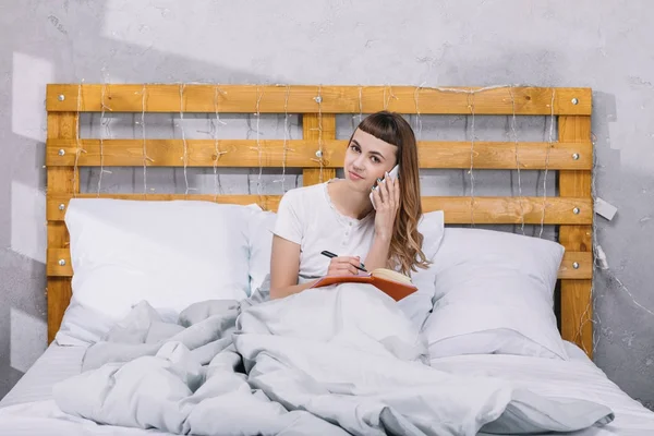 Lächelndes Mädchen, das etwas ins Notizbuch schreibt und im Bett mit dem Smartphone spricht — Stockfoto