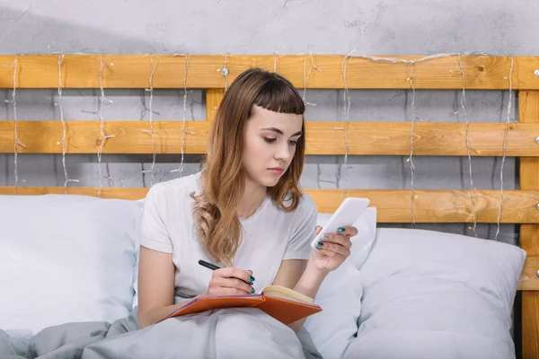 Girl writing something to notebook and looking at smartphone in bed — Stock Photo