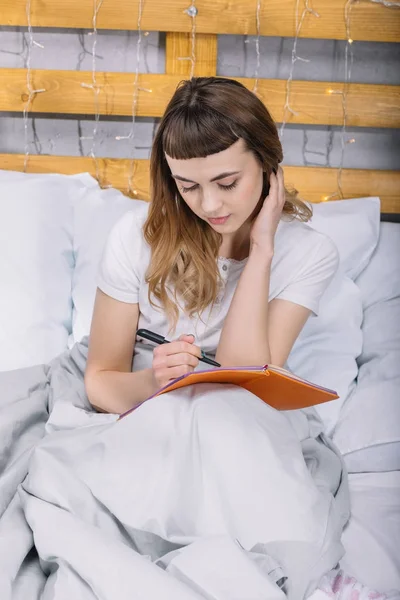 Chica escribiendo algo a cuaderno en la cama por la mañana - foto de stock