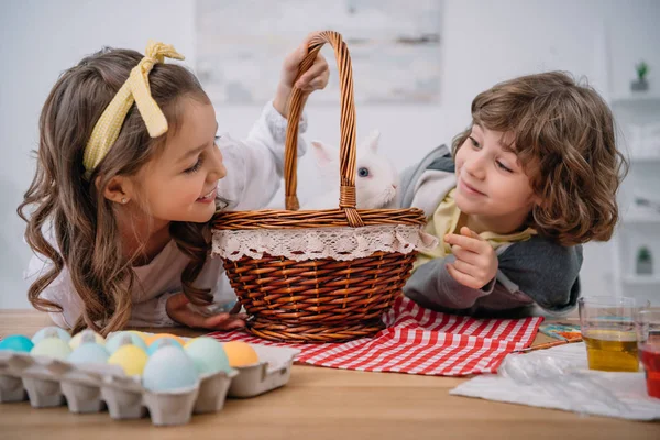 Niños pequeños y felices jugando con conejo lindo en la cesta en la mesa con huevos de Pascua - foto de stock