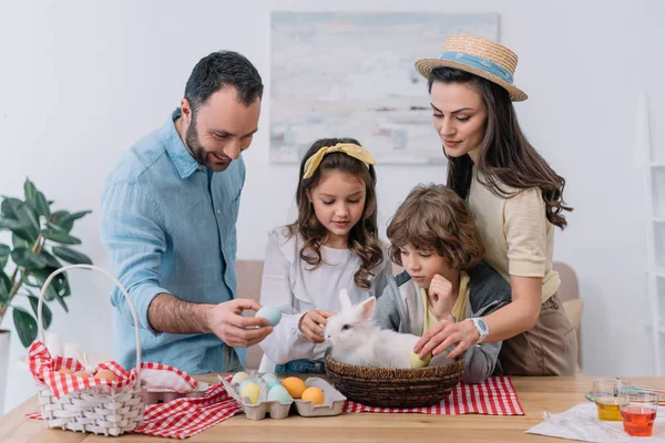 Giovane famiglia che si prepara per le vacanze pasquali con coniglietto seduto nel cesto sul tavolo — Foto stock