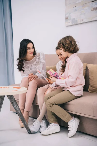 Niños sentados con la madre en el sofá y mostrando sus tarjetas de felicitación para el día de la madre - foto de stock