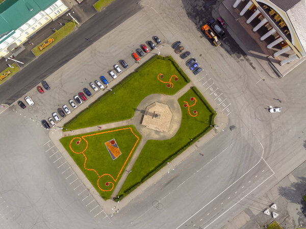 City square with geometric flowerbed patterns, monument and car park, aerial