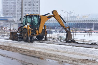 Rostov-on-Don / Russia - January 2018:  A yellow excavator in a snowfall digs a pit on the roadside in a town with warning signs clipart