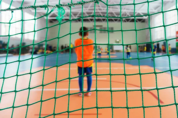 Young football players in bright uniforms play football in a closed sports complex behind a football gate with a close-up grid